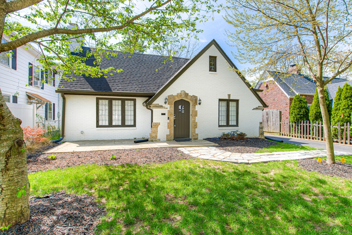 The home is framed by a lush green lawn and features a stone pathway leading to the front entrance. Brightly colored landscaping adds to the inviting exterior, highlighted by large windows on either side of the door, while trees offer gentle shade.