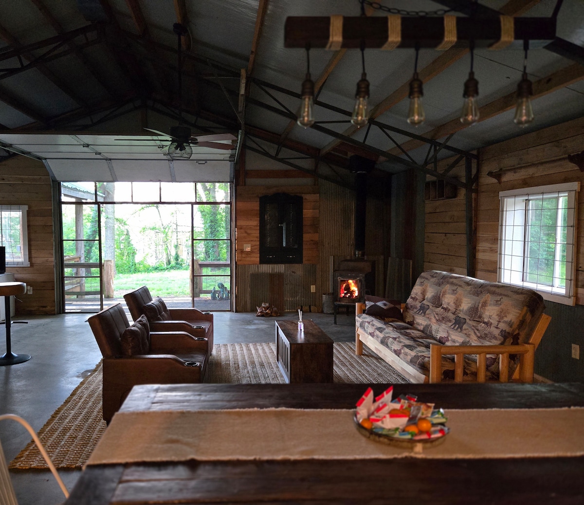An inviting living area features a cozy couch adorned with nature-themed upholstery and two wooden armchairs. A rustic coffee table sits in the center, surrounded by a woven rug. Natural light pours in through large windows, illuminating the space, while a small fireplace provides a warm focal point.