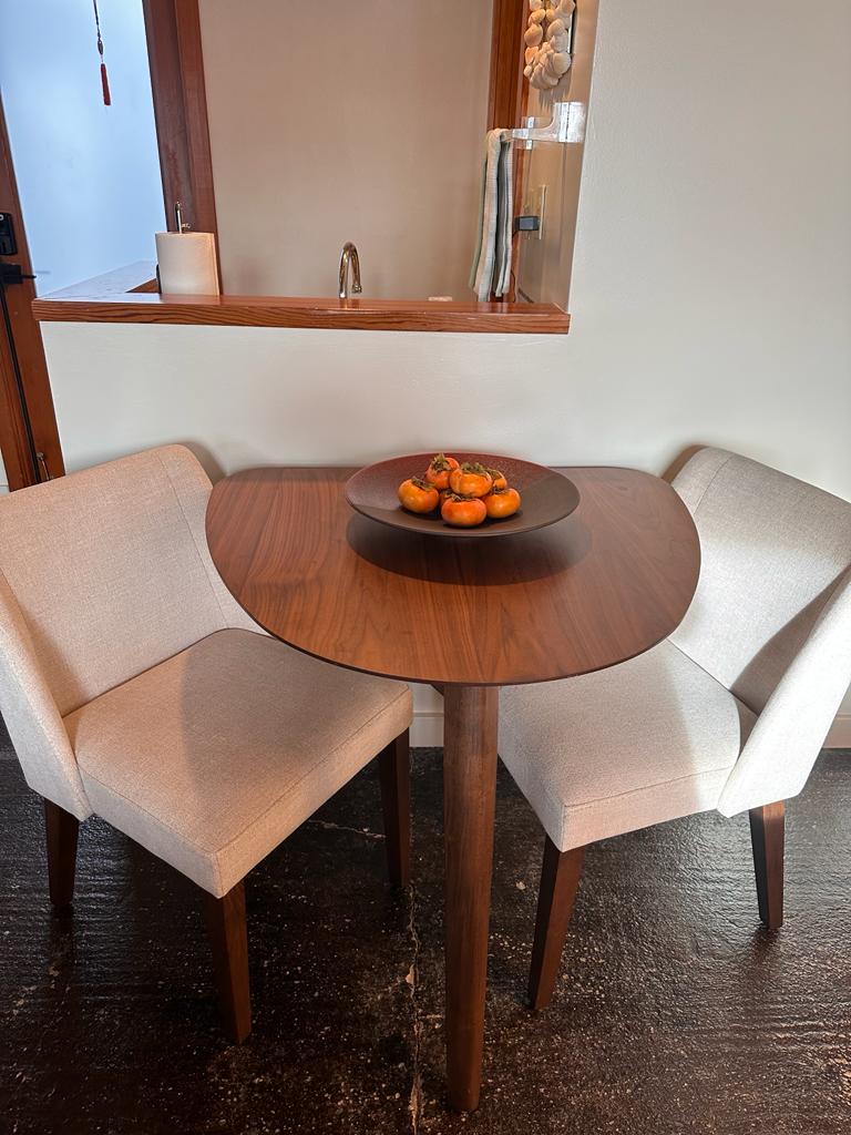 A small, round wooden table is paired with two upholstered chairs. The table is centered with a bowl of oranges, providing a welcoming touch. A small counter space with a modern faucet is visible in the background, adding to the functional atmosphere.