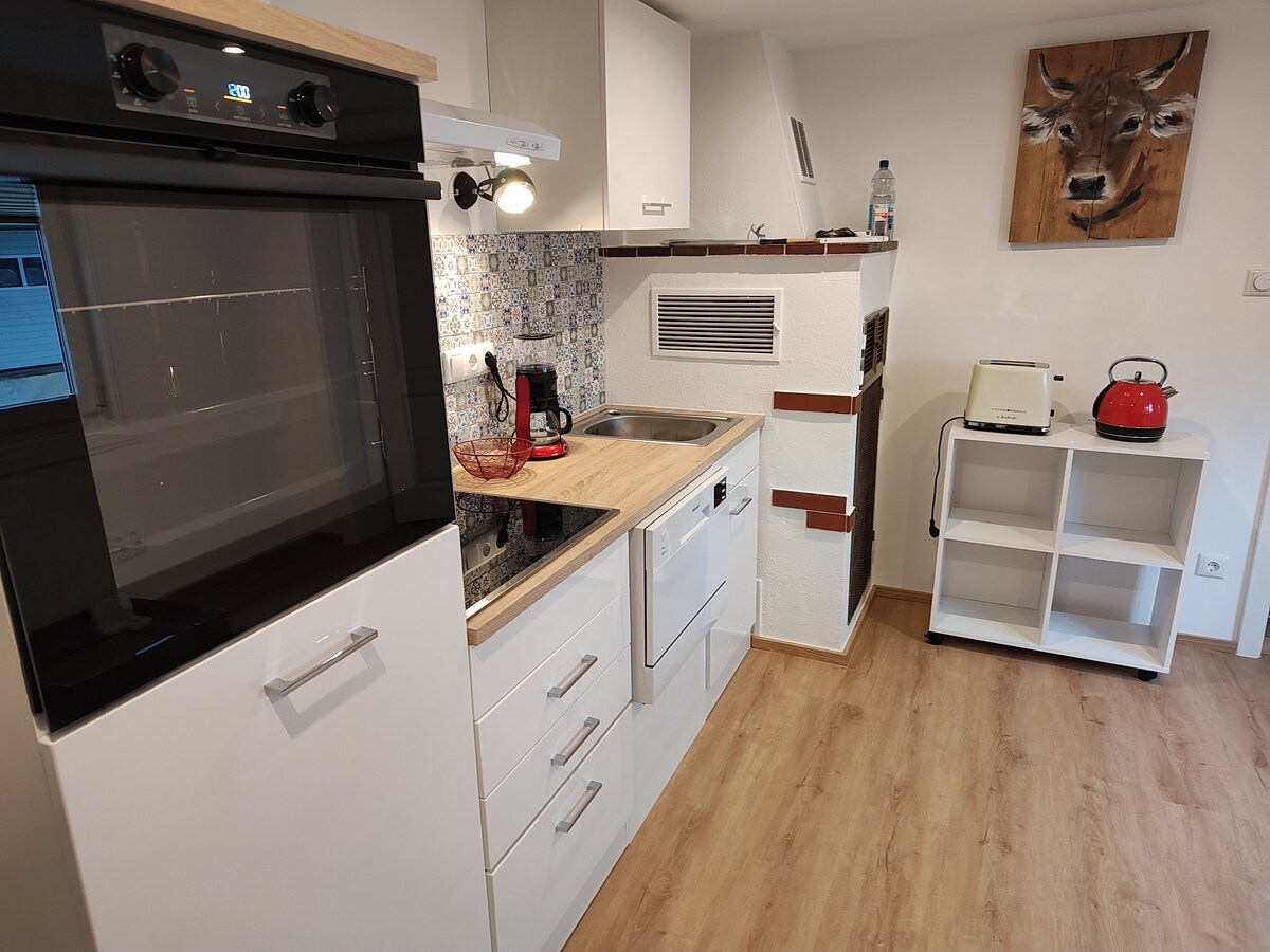The modern kitchen features sleek white cabinets and a wooden countertop. Essential appliances include an oven, a stovetop, a refrigerator, and a dishwasher, arranged efficiently. Decorative tiles behind the sink add a touch of character, while a small dining table and a kettle complete the space.