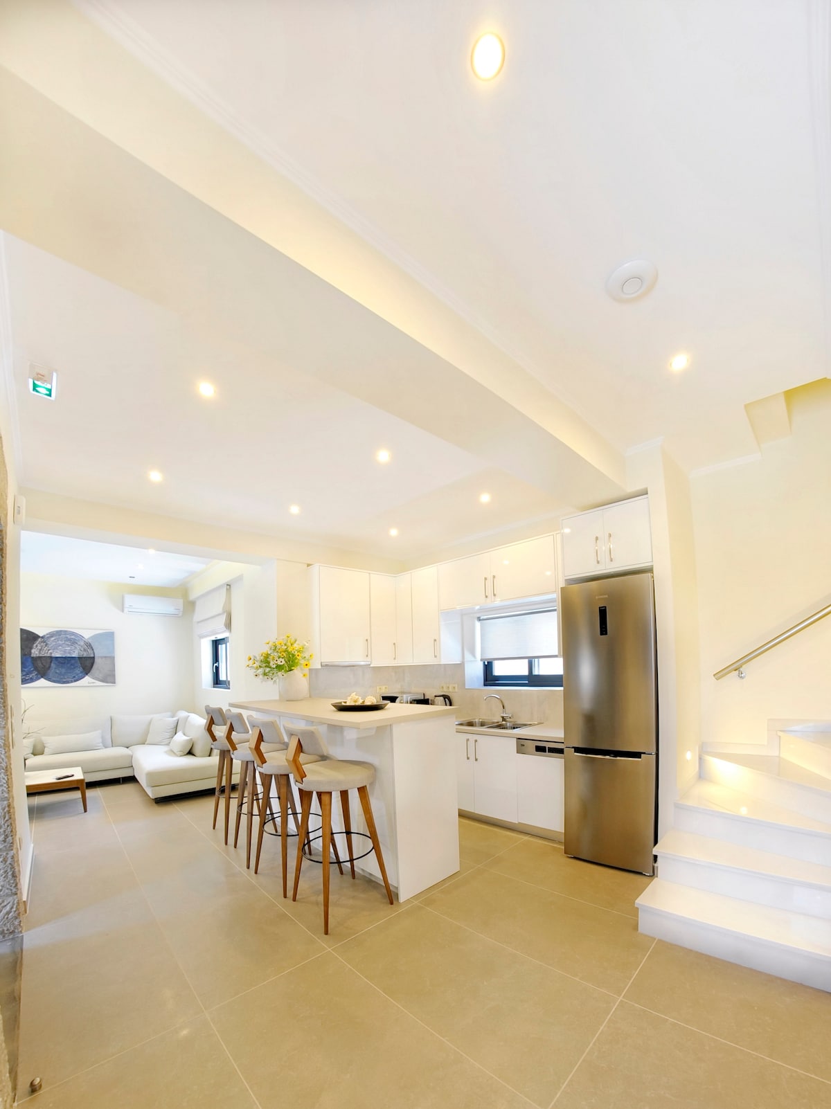 An open-plan kitchen and living area is visible, featuring light-colored cabinetry and stainless-steel appliances. Four wooden barstools line the kitchen island, and a comfortable seating arrangement with soft cushions is situated nearby. Natural light fills the space through large windows, creating an airy ambiance.
