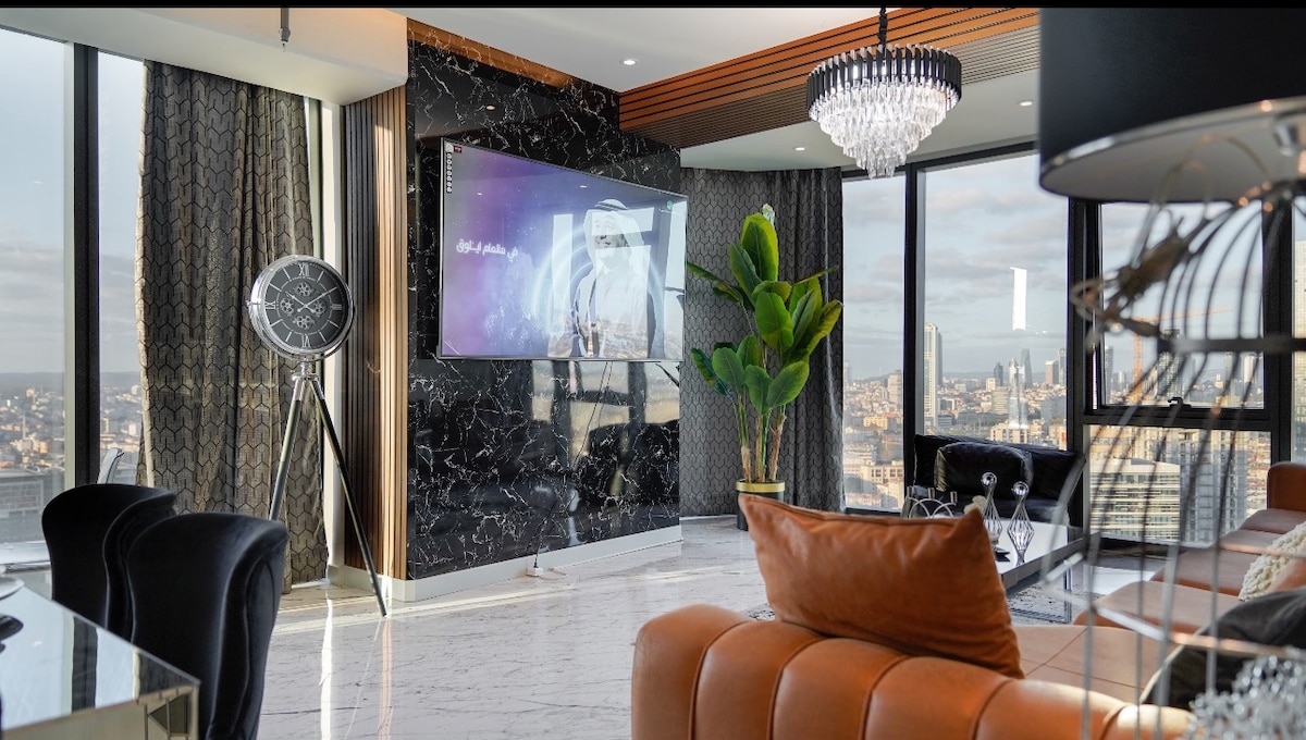 A modern living area is showcased, featuring a large screen television mounted on a decorative wall with dark marble. Plush seating in deep colors surrounds a glass table, while a tall plant and an eye-catching chandelier add elegance. Floor-to-ceiling windows highlight the stunning city view.