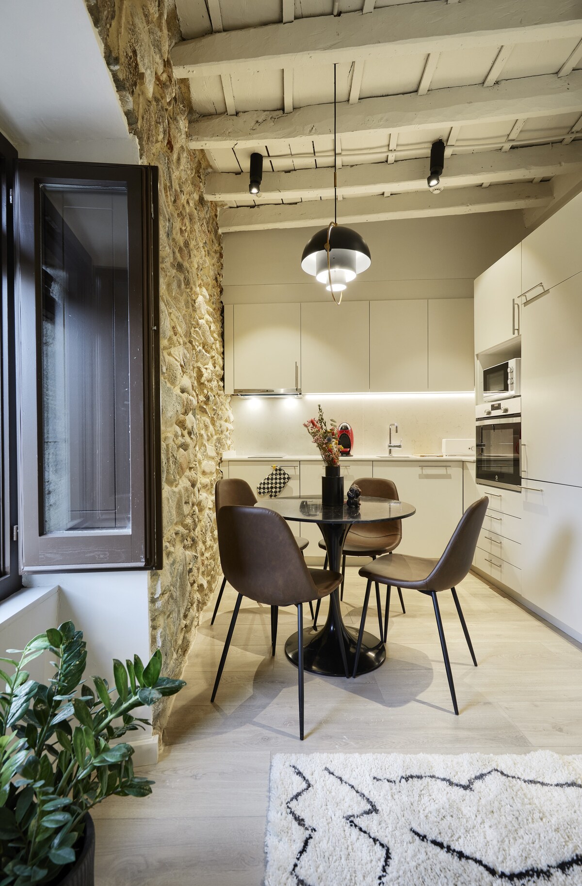 A modern kitchen area features a round black dining table surrounded by four brown chairs. A stone wall complements sleek cabinetry and white countertops. Bright overhead lighting enhances the space, while a window allows natural light to illuminate the room.