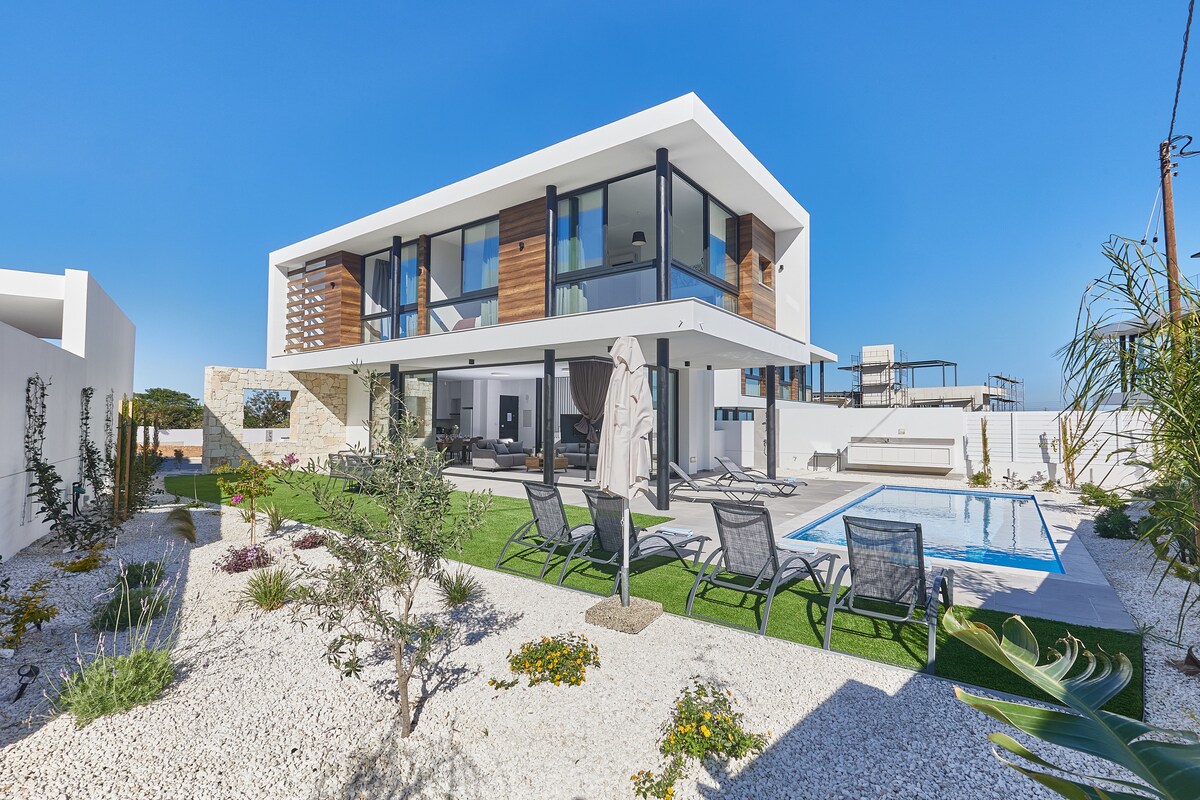 The contemporary two-story villa is set within a landscaped garden featuring an outdoor pool. A spacious patio with reclining sun loungers is visible, alongside areas of greenery and decorative plants. Large glass panels provide natural light to the upper-level balconies and enhance the modern aesthetic.