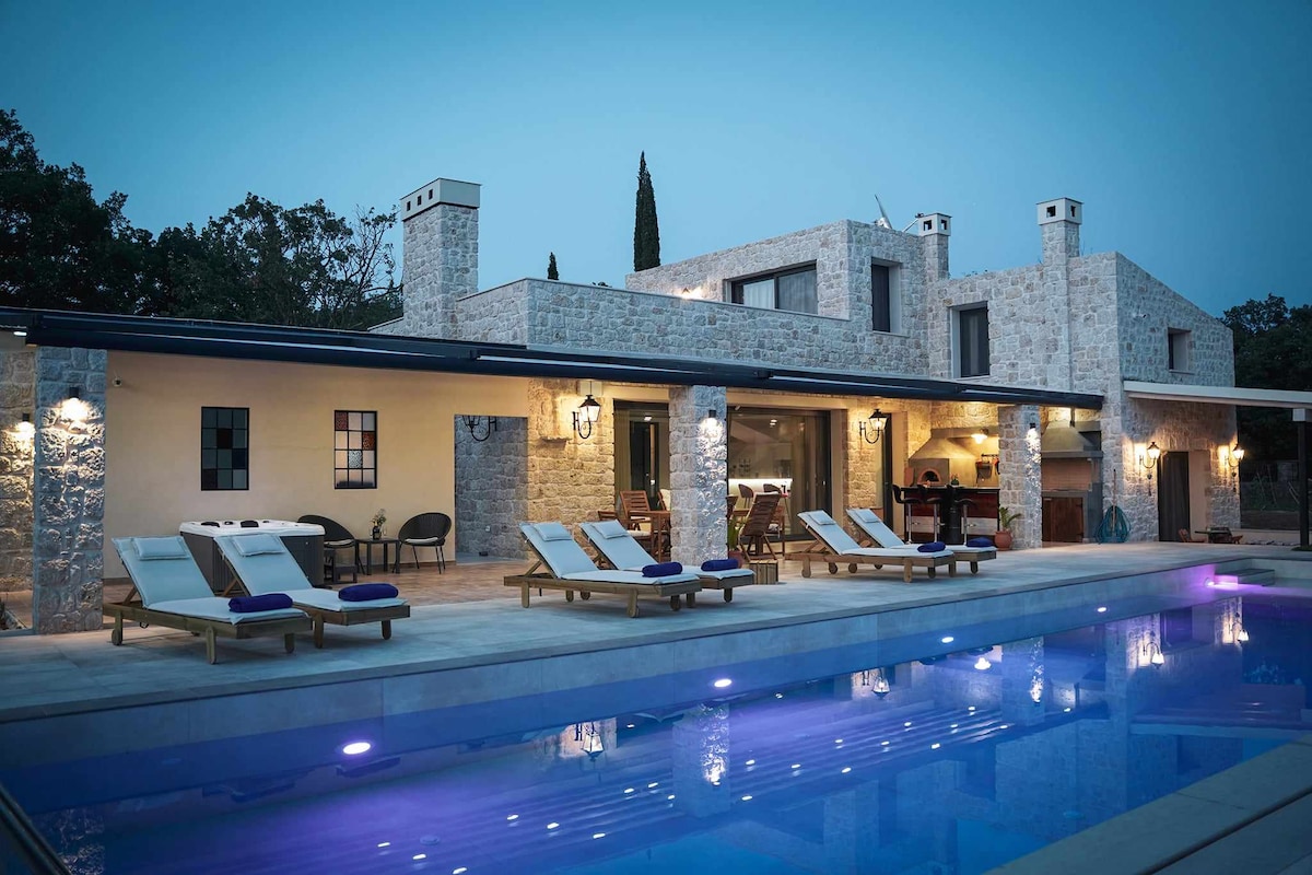 The exterior of a newly built stone villa is captured at dusk, featuring a large swimming pool illuminated in soft blue light. Sun loungers are arranged nearby on a patio, with a cozy outdoor seating area visible through expansive glass doors.