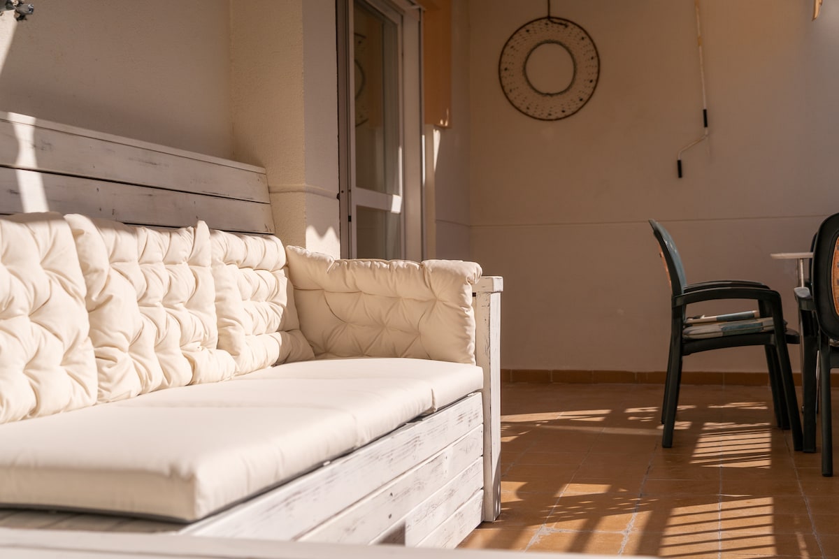 A spacious terrace features a light-colored sofa with plush cushions, providing a comfortable seating area. A round clock is mounted on the wall, and a dining table with chairs is visible, enhancing the outdoor space's functionality.