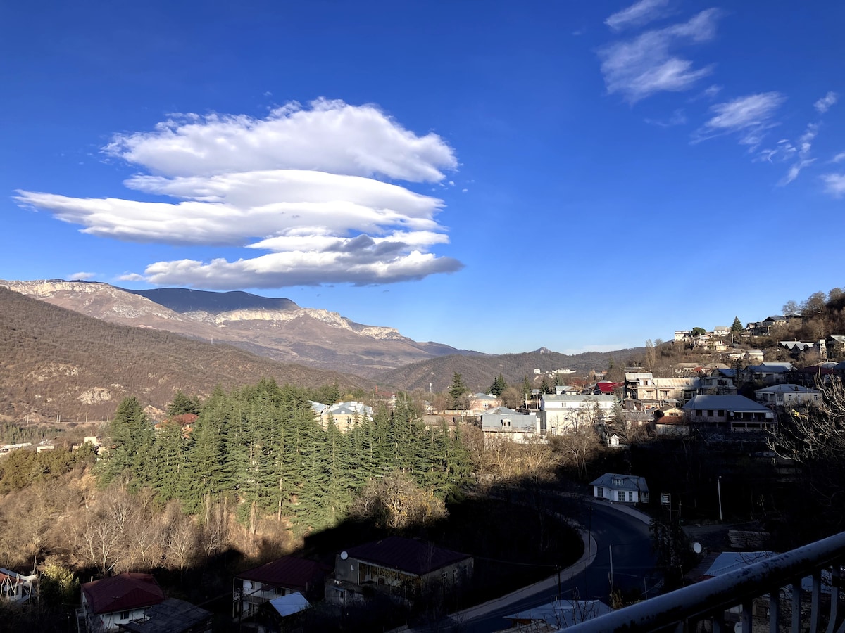 A scenic landscape showcases rolling mountains against a clear blue sky. Soft, layered clouds hover above the horizon, while a lush forest of evergreen trees fills the foreground. A peaceful neighborhood with scattered homes is nestled among the slopes, creating a serene atmosphere.