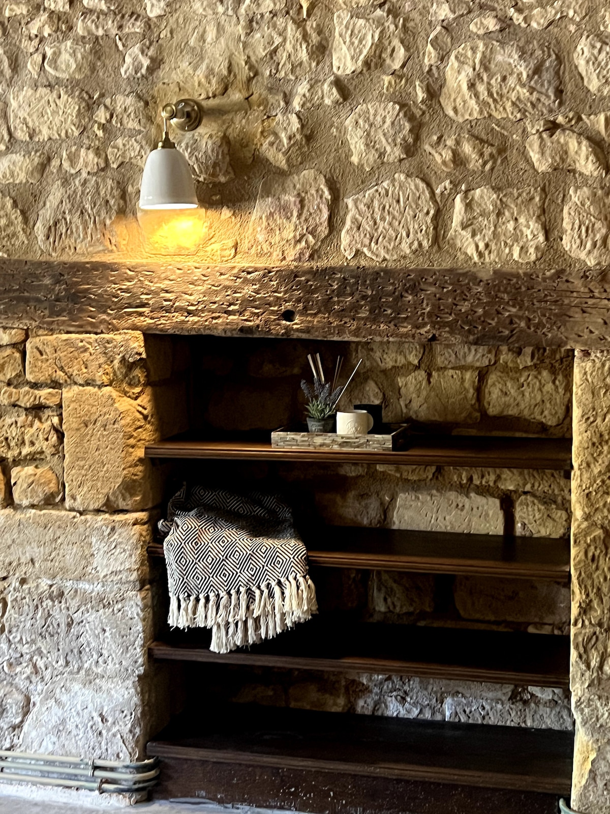 A rustic shelving unit is set against a stone wall, illuminated by a warm wall-mounted light. The shelves display a small decorative arrangement with a plant and a cup, alongside a cozy woven blanket draped partially over the shelf.