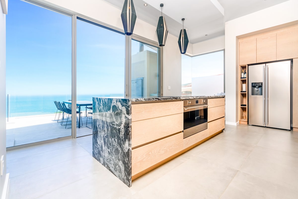A modern kitchen space features a large marble island with an integrated sink and sleek cabinetry. Floor-to-ceiling glass doors provide stunning ocean views and natural light, while contemporary pendant lights hang overhead, adding elegance to the cooking area.