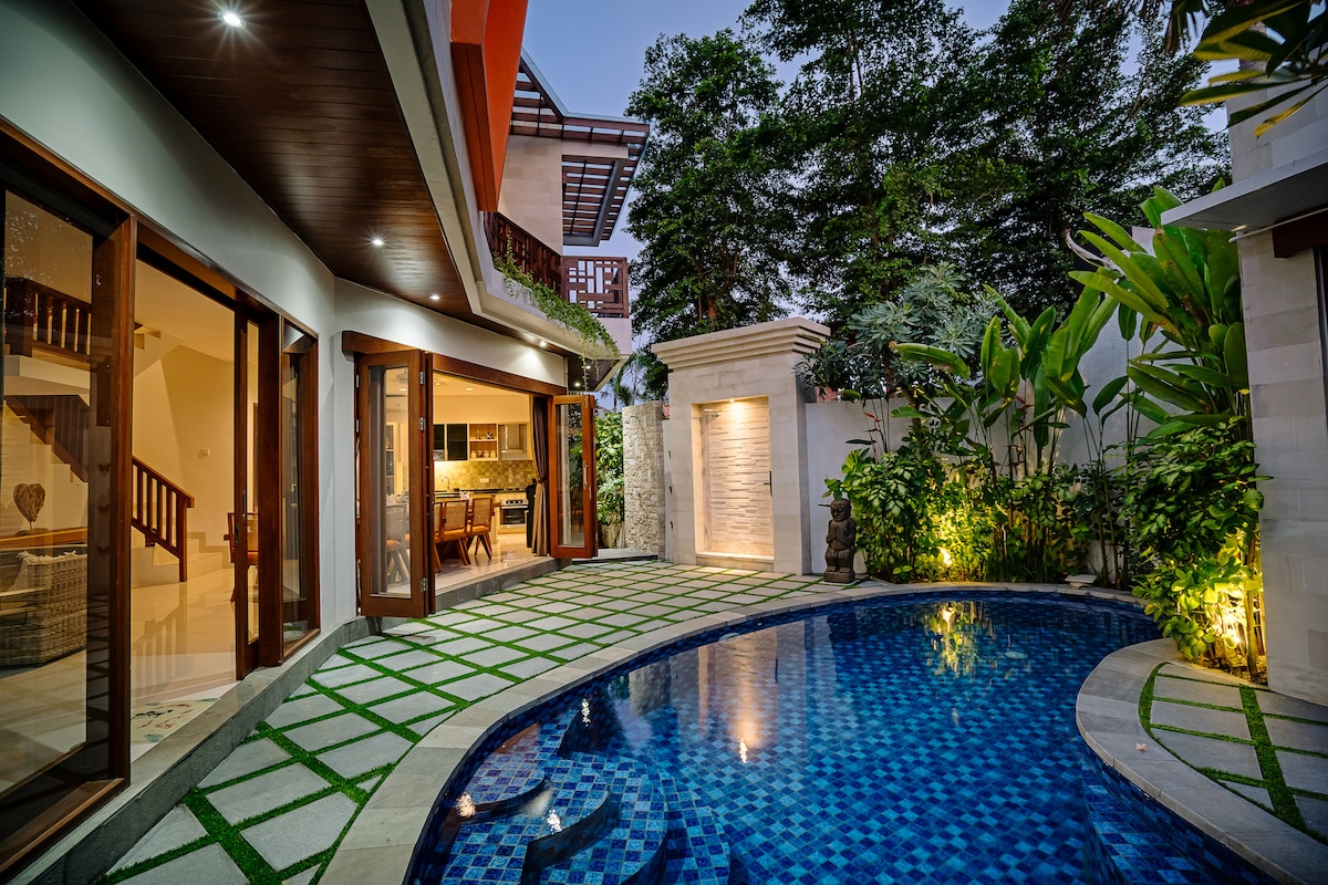A serene courtyard showcases a private swimming pool with blue mosaic tiles. The pool area is framed by lush greenery and elegantly designed landscaping. Large glass doors connect the outdoor space to the living area, enhancing the indoor-outdoor flow of the villa.