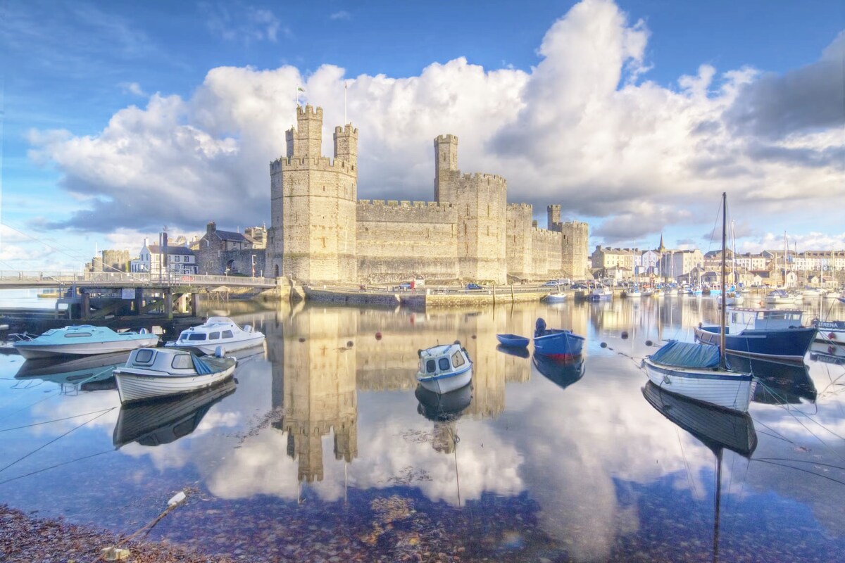 The historic Caernarfon Castle stands majestically by the water, surrounded by boats gently bobbing on the calm surface. Reflections of the castle and fluffy clouds are visible in the still water, creating a serene backdrop against the vibrant blue sky.