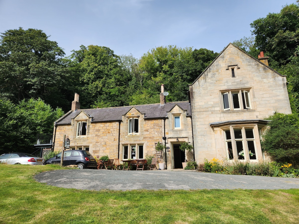 A charming stone house is set amidst lush greenery, showcasing large windows that allow natural light to fill the interior. An inviting outdoor area features a circular driveway and a dining table, surrounded by flowering plants. The structure combines rustic and elegant architectural elements.