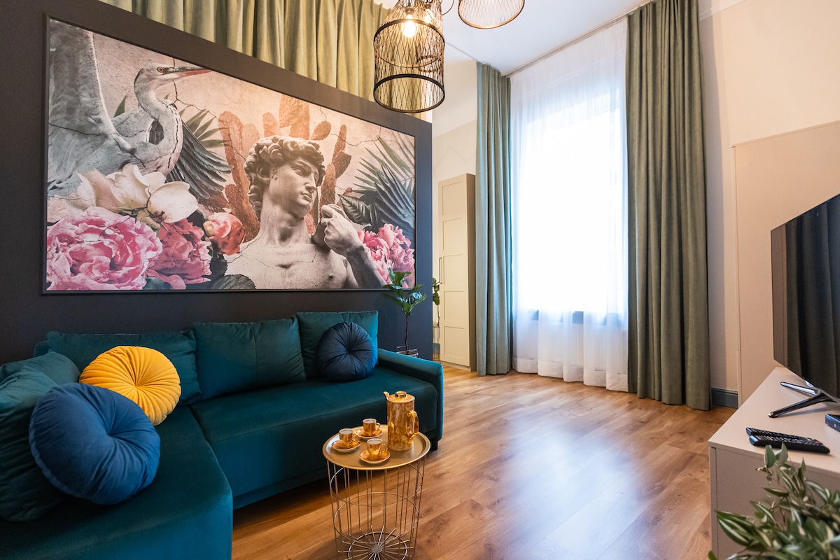 A stylish living area showcases a deep green sectional sofa adorned with a yellow cushion. A round coffee table displays refreshments, while a large artwork above the sofa features classical figures framed by floral elements. Natural light fills the space through tall windows, dressed with sheer curtains.