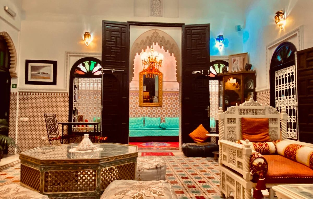 An elegant seating area features intricate Moroccan decor, with a low table surrounded by plush sofas and colorful cushions. Traditional arches are adorned with ornate details, and vibrant stained glass windows provide a soft glow, enhancing the inviting ambiance of the space.