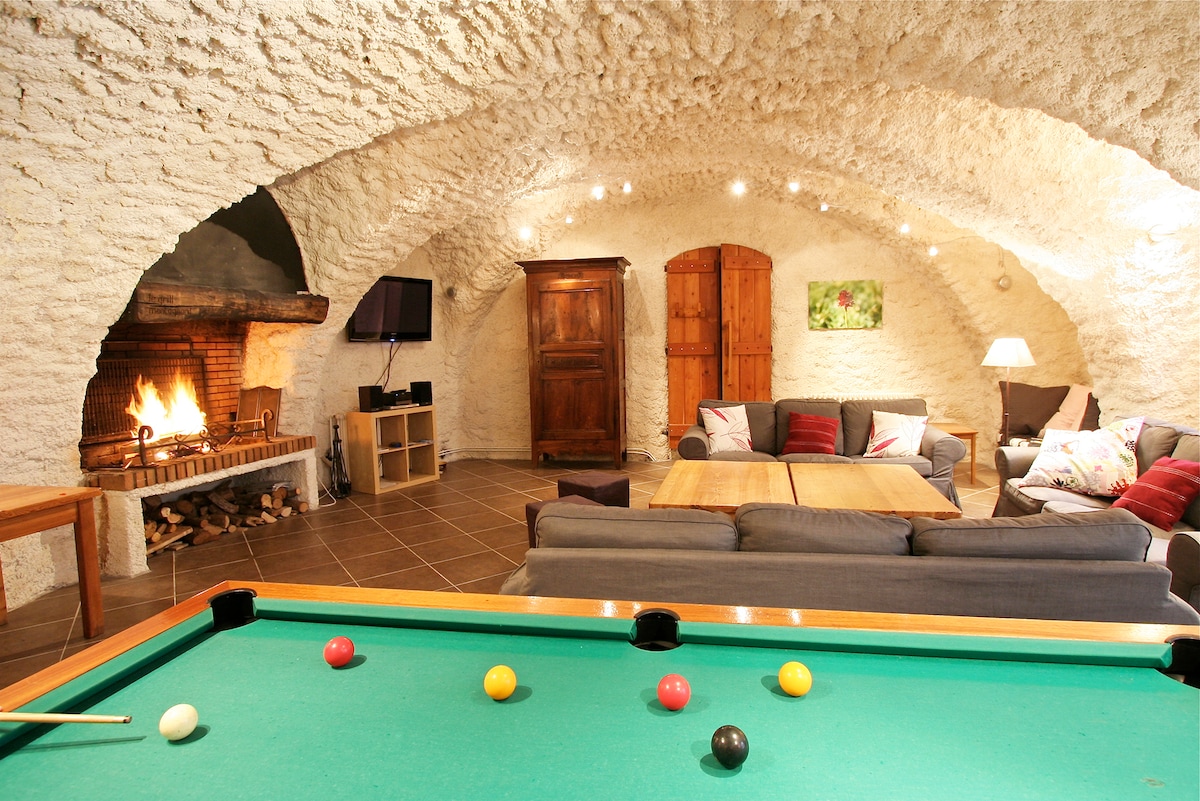 A spacious vaulted living area features a billiard table, comfortable sofas, and a cozy fireplace. Warm light from recessed ceiling fixtures enhances the textured walls, while wooden furniture adds to the inviting atmosphere. A television and storage cabinet are positioned against the wall.