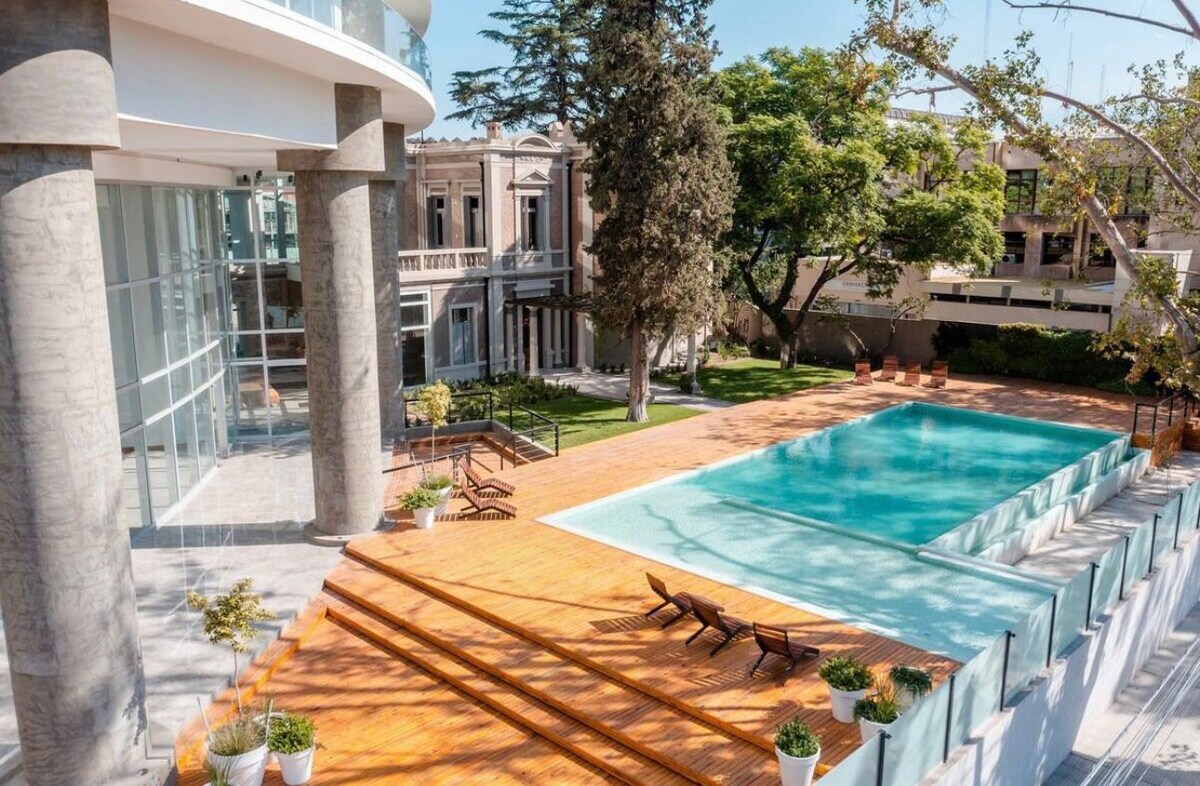 An outdoor swimming pool is surrounded by a wooden deck and lush greenery. Sun loungers are placed along the side of the pool, and large windows of the building reflect the sunny atmosphere. The area exudes a peaceful ambiance suitable for relaxation.