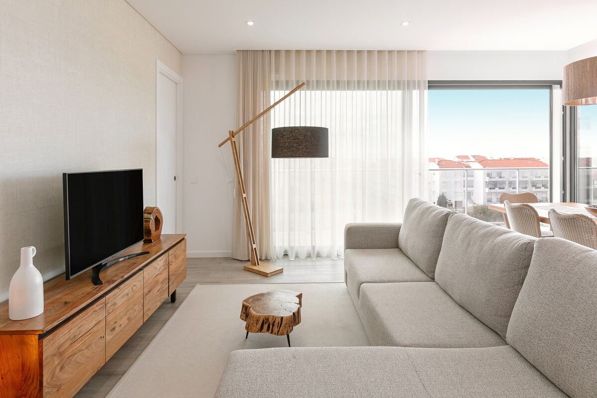 A spacious living room features a comfortable sofa and a wooden coffee table. Natural light fills the area through large glass doors, which lead to a balcony. A sleek television is positioned on a wooden console, complemented by soft lighting from a stylish floor lamp.