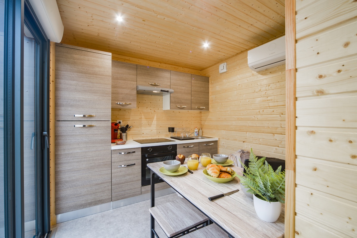 The kitchen area features contemporary cabinetry and modern appliances. A dining table is set with plates and fresh pastries, complemented by a potted plant. Natural light streams in through large glass doors, enhancing the inviting atmosphere of this functional space.