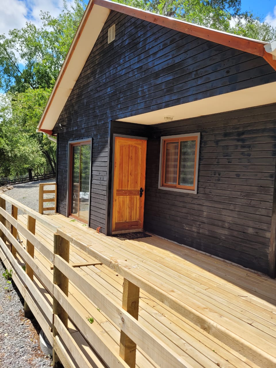 A welcoming entrance is presented with a wooden door framed by two windows. A spacious deck made of light wood extends from the entrance, surrounded by greenery, inviting guests to relax outdoors.