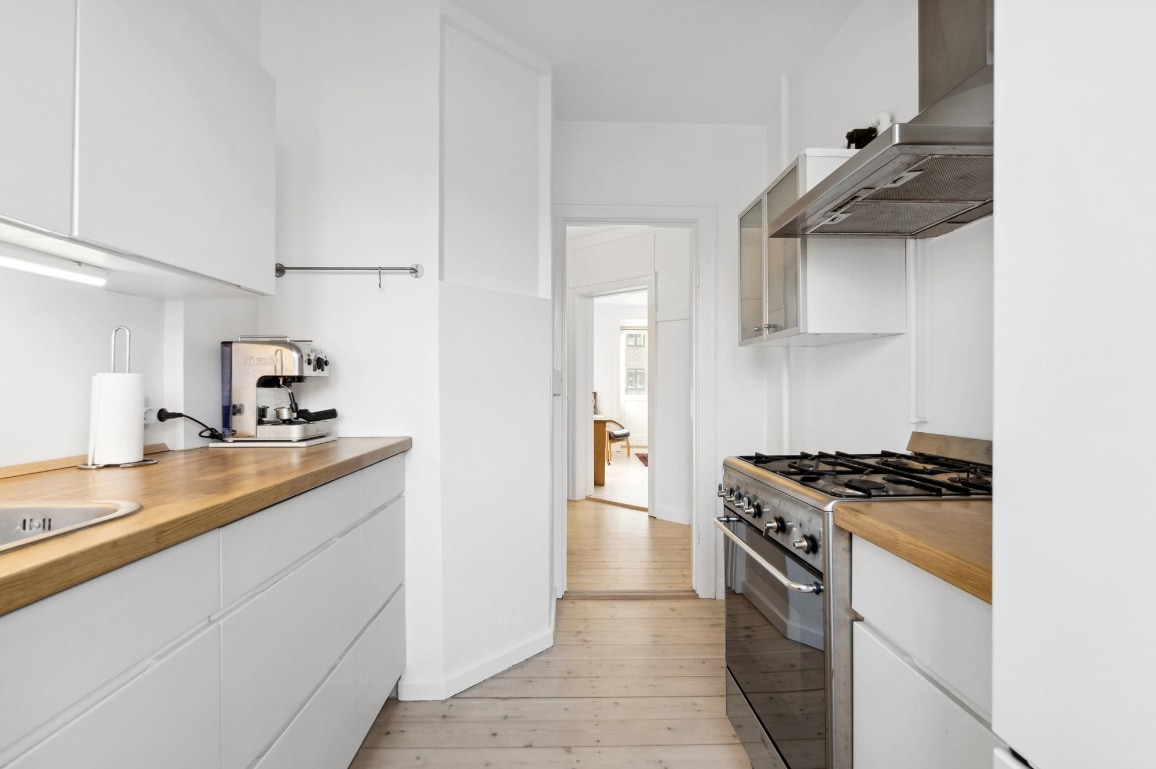 A clean and modern kitchen features white cabinetry with wooden countertops. A coffee machine and sink are visible on the left side, complemented by a gas stove on the right. The space is well-lit with natural light, creating an inviting atmosphere.