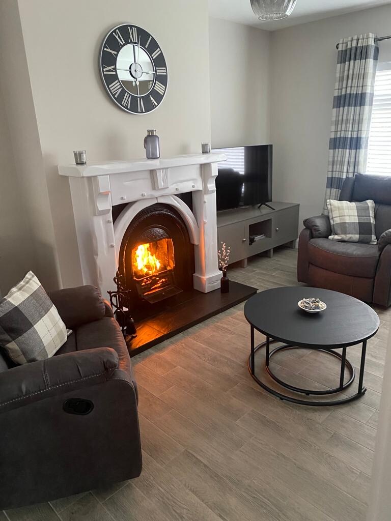 A cozy living area features a traditional fireplace with a glowing fire. Two comfortable armchairs are positioned nearby, with soft cushions adding detail. A round coffee table sits centrally, while a modern television is mounted on a wall opposite, complemented by patterned curtains.