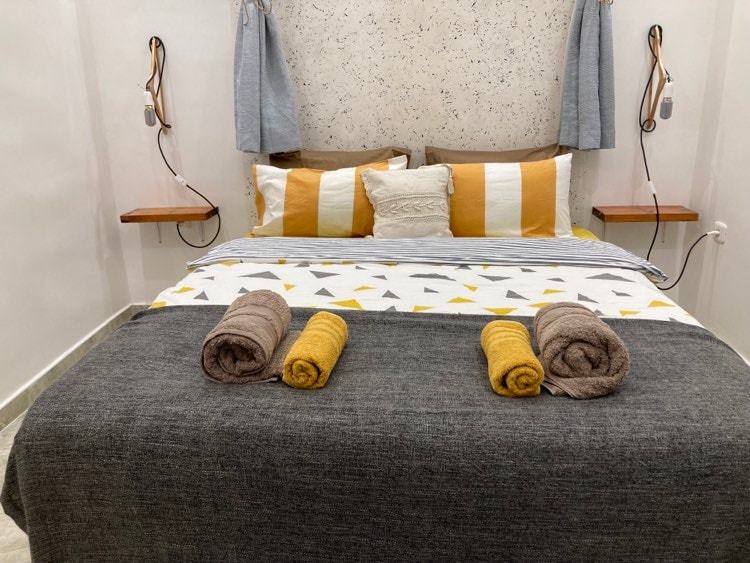 A well-organized sleeping area features a spacious bed dressed in a patterned blanket. Fluffy towels in shades of gray and yellow are neatly arranged at the foot of the bed. Soft lighting is provided by wall-mounted lamps on either side.