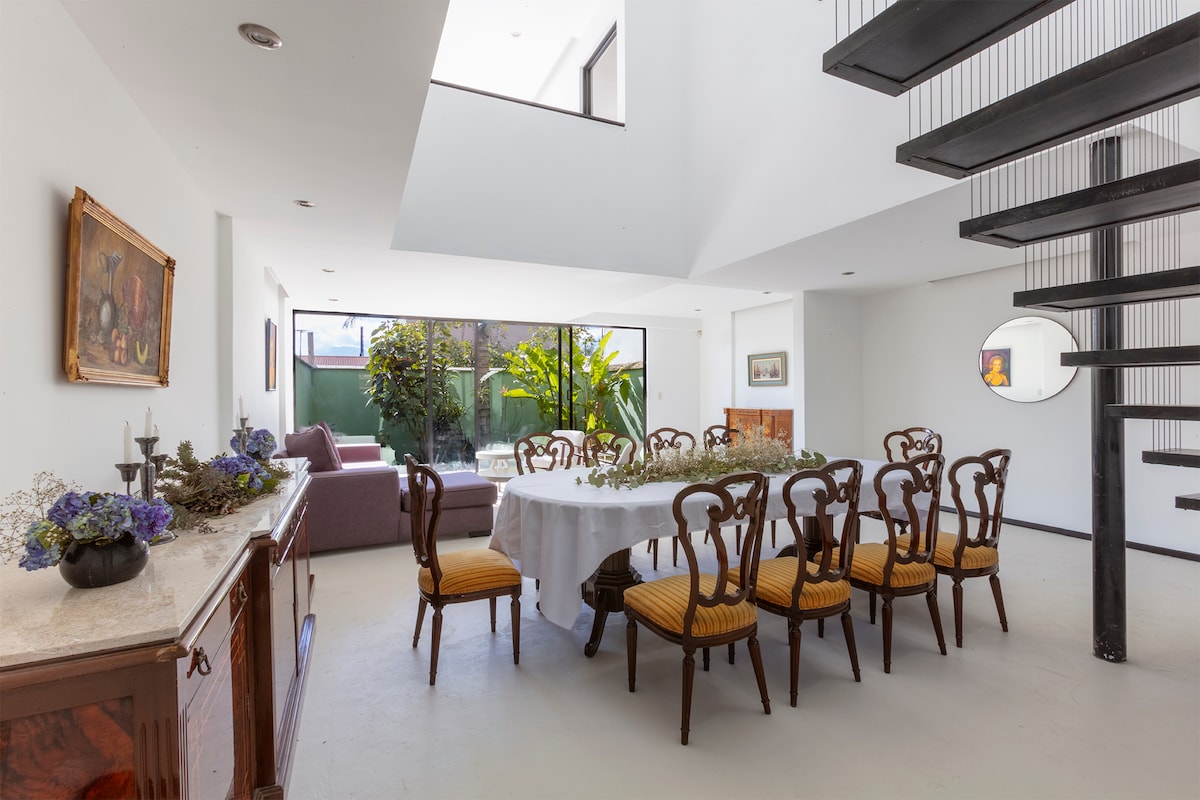 A spacious dining area is highlighted by a large table surrounded by classic wooden chairs. Large windows provide natural light and views of greenery. A modern spiral staircase leads to the upper level, and a cozy seating area is visible in the background.