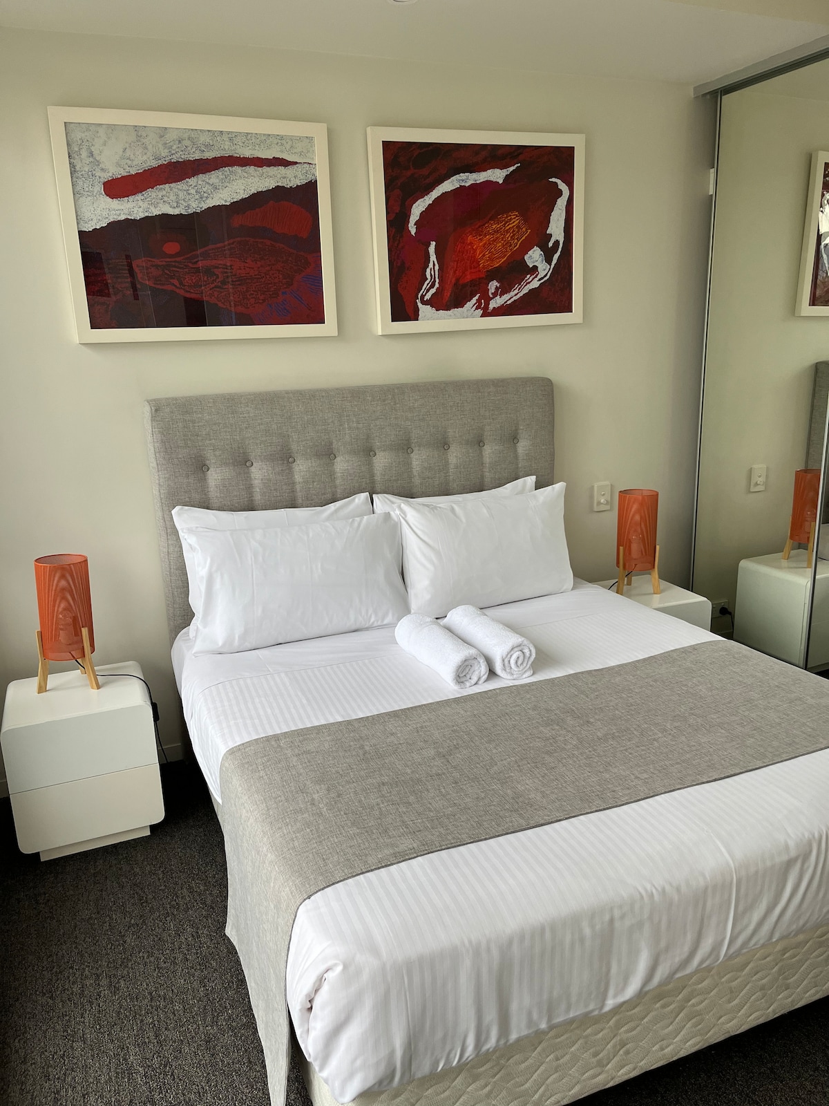 A neatly arranged bedroom features a spacious bed with crisp white linens and a light gray throw blanket. Two decorative towels are placed at the foot of the bed. Framed artwork adorns the wall above the bed, and mirrored closet doors reflect the serene setting.