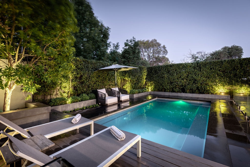 A well-maintained swimming pool is surrounded by neatly trimmed greenery, creating a serene atmosphere. Lounge chairs are positioned alongside the pool, complemented by a shaded seating area under an umbrella. Soft lighting enhances the ambiance, inviting relaxation during evening hours.