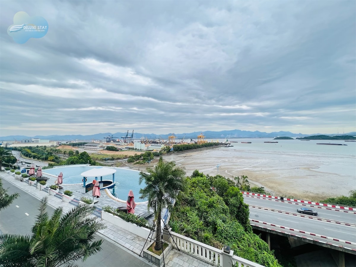 A serene waterfront view features distant mountains and a calm sea. The foreground includes a stylish infinity pool bordered by palm trees, while a coastal road runs alongside. Umbrellas provide shade for outdoor seating areas, inviting relaxation amid nature.