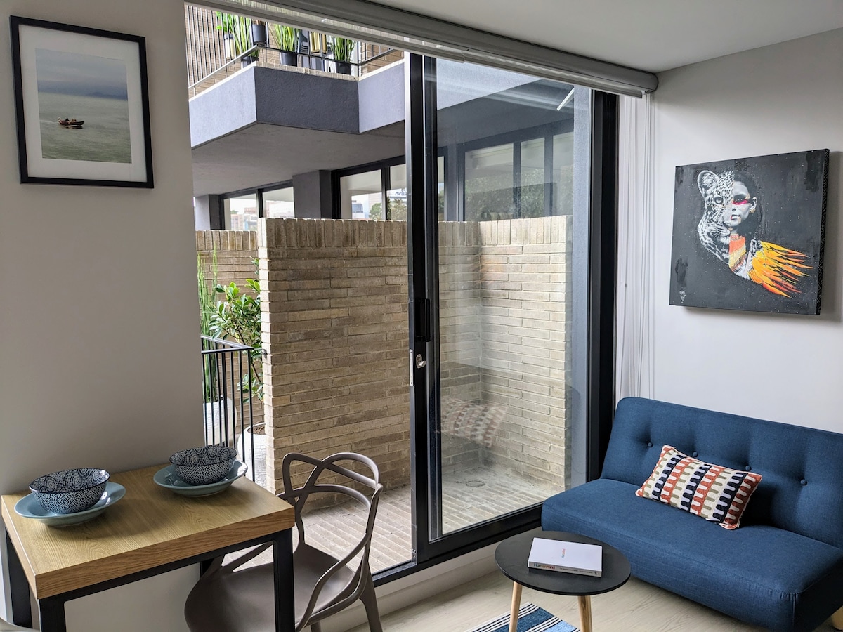 A bright corner of the apartment showcases a blue sofa paired with a small wooden dining table, set with two bowls. Large glass doors open onto a private outdoor space, framed by brick walls. Art pieces adorn the walls, adding a touch of character.