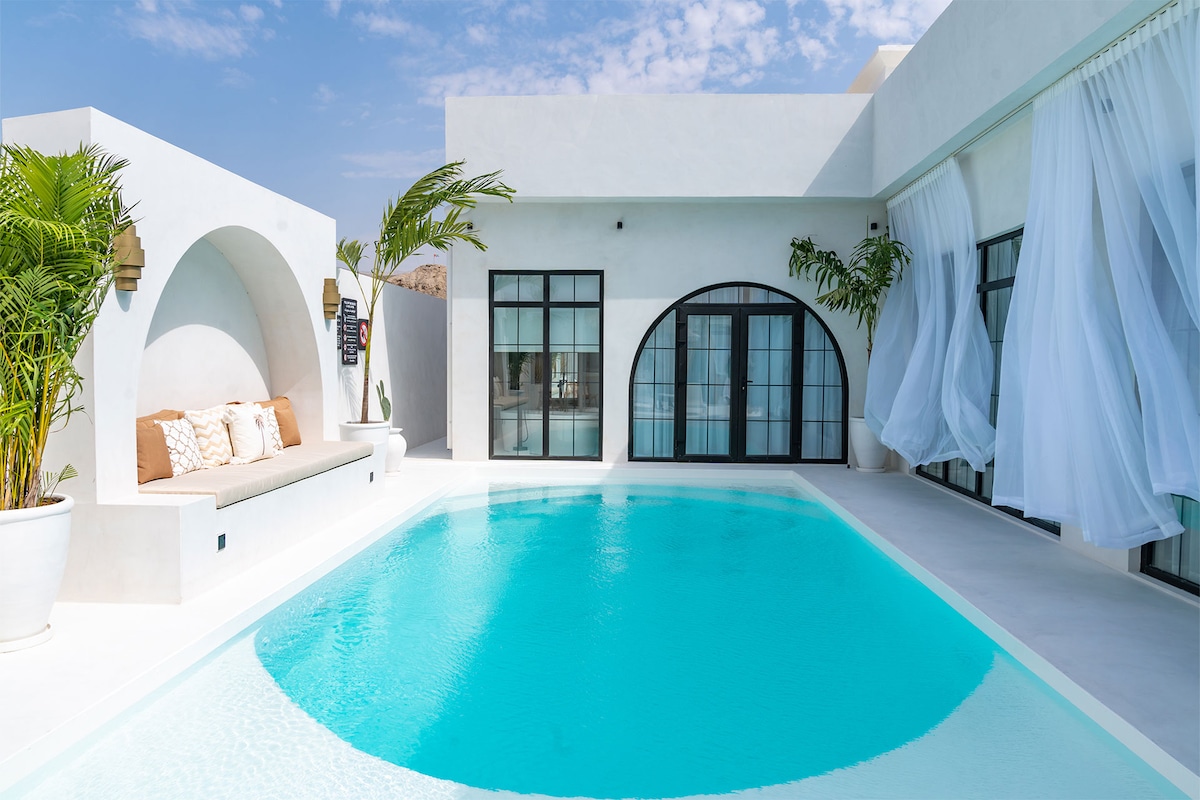 A luxurious pool area features a clear blue swimming pool surrounded by white walls and modern architecture. An outdoor seating area with cushions is visible under an arch, while large glass doors provide views of the interior. Flowing sheer curtains enhance the space.