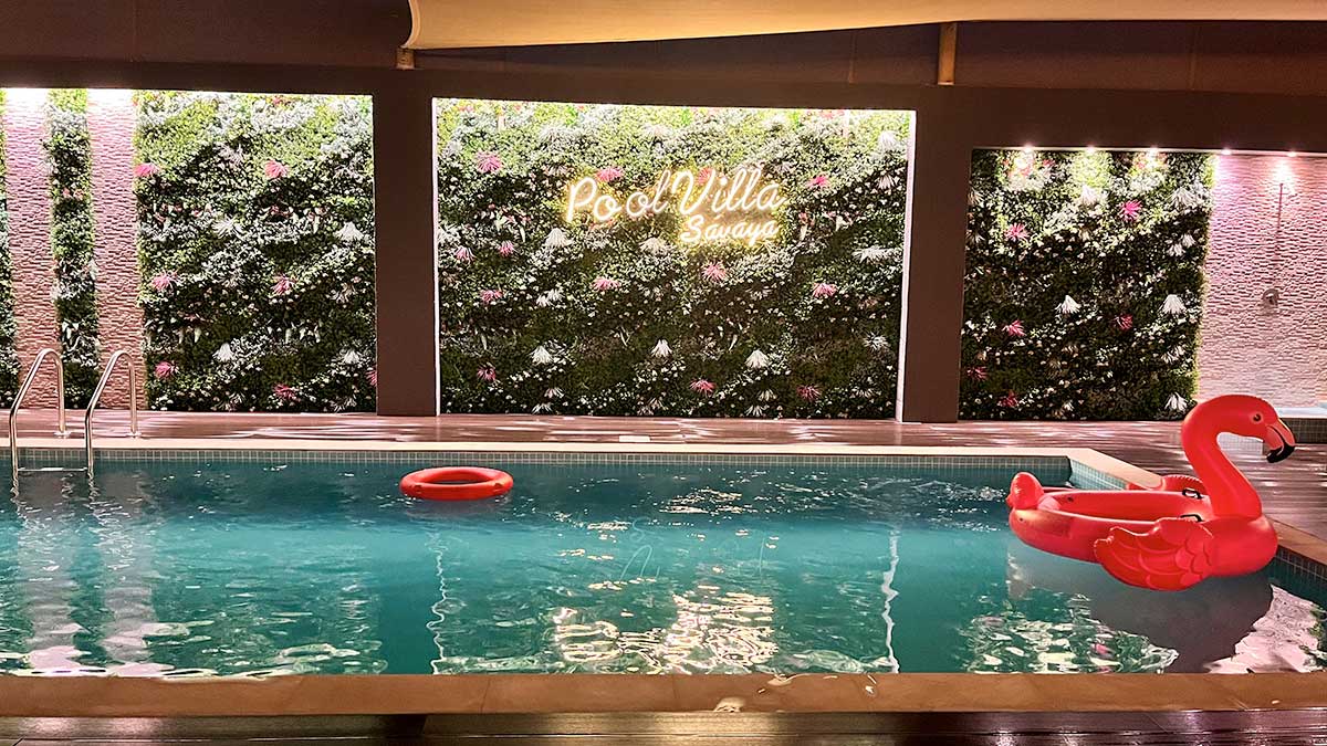 A serene indoor pool area is highlighted by a lush green wall adorned with flowers. A red float in the shape of a flamingo is positioned in the water, and soft ambient lighting creates a relaxing atmosphere around the space.