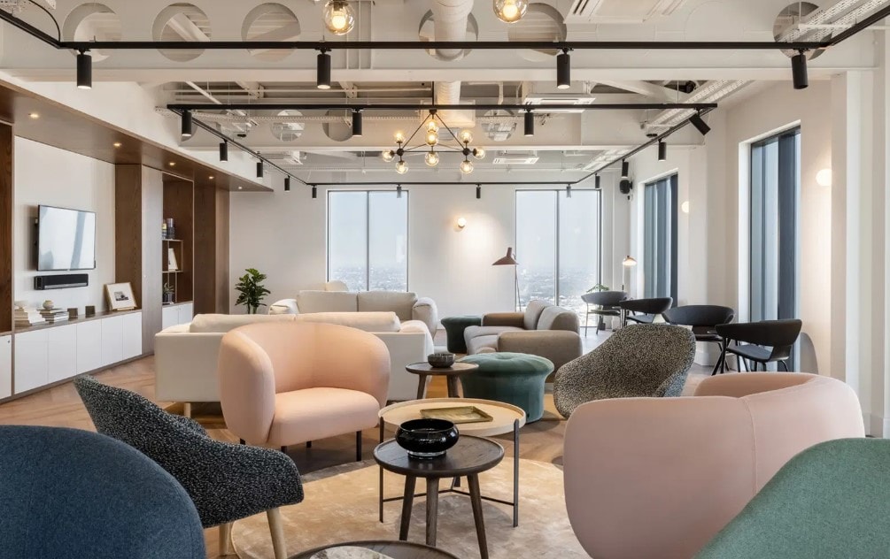A modern co-working space features various seating arrangements with soft, pastel-toned chairs alongside dark-colored ones. Large windows allow ample natural light and offer views of the city. Coffee tables with small decorative items are scattered throughout the area, creating an inviting atmosphere.