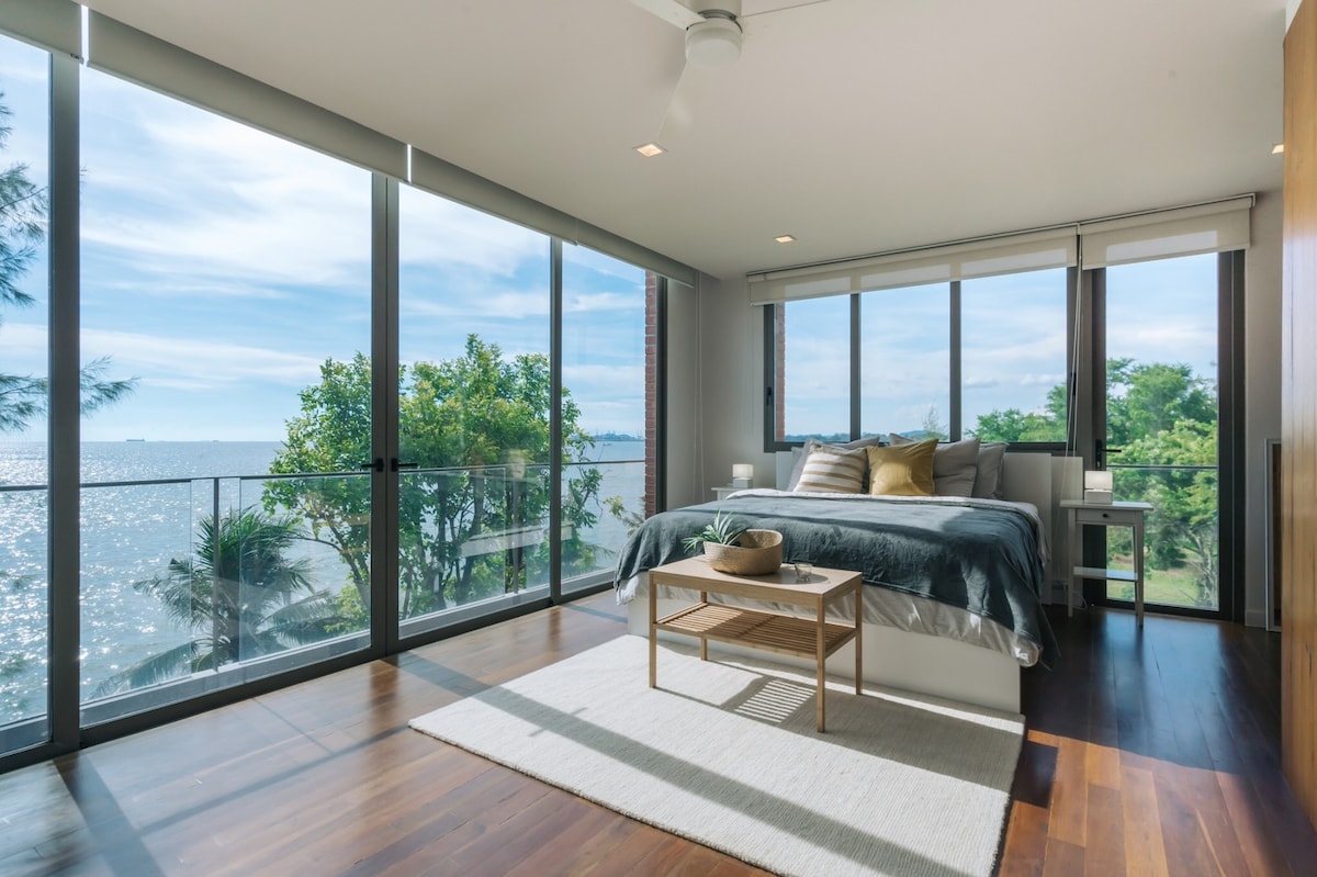 A serene bedroom features a spacious bed dressed in neutral tones, with large glass windows offering unobstructed sea views. Natural light floods the room, illuminating the hardwood floor and a simple area rug. A tasteful arrangement of decor enhances the tranquil atmosphere.