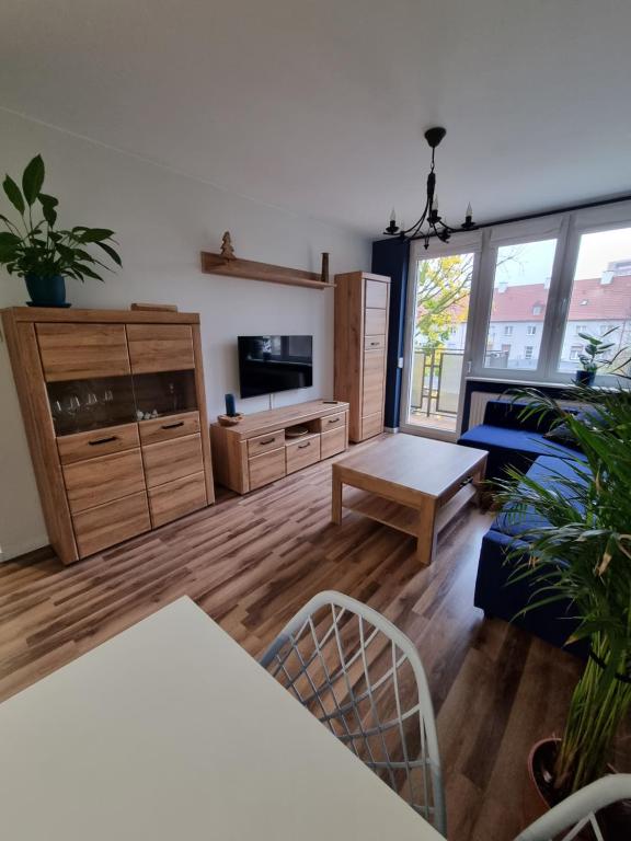 A living area features a warm wooden cabinet and a media console, complemented by a coffee table. Indoor plants are arranged throughout the space. Natural light enters from large windows, enhancing the cozy atmosphere created by blue furnishings and wooden flooring.