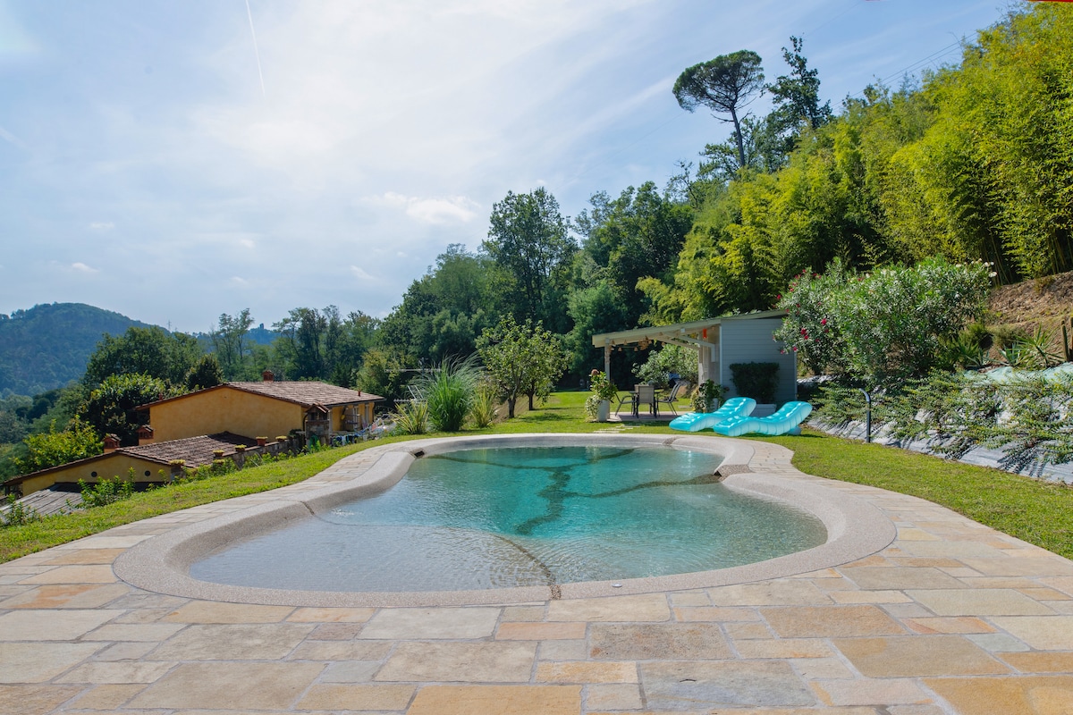 A serene infinity pool is set against a backdrop of lush greenery and rolling hills. Sun loungers are positioned nearby on a tiled terrace, providing a perfect spot for relaxation. The landscape is dotted with trees and flowering plants, enhancing the natural beauty.