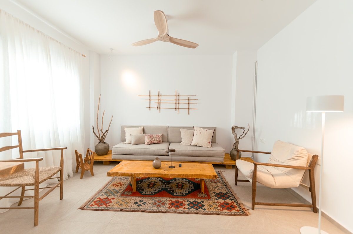A modern living area is presented, featuring a neutral grey sofa adorned with cushions. A wooden coffee table sits on a decorative area rug, surrounded by a mix of wooden chairs. Natural light filters through sheer curtains, creating a bright and inviting space.