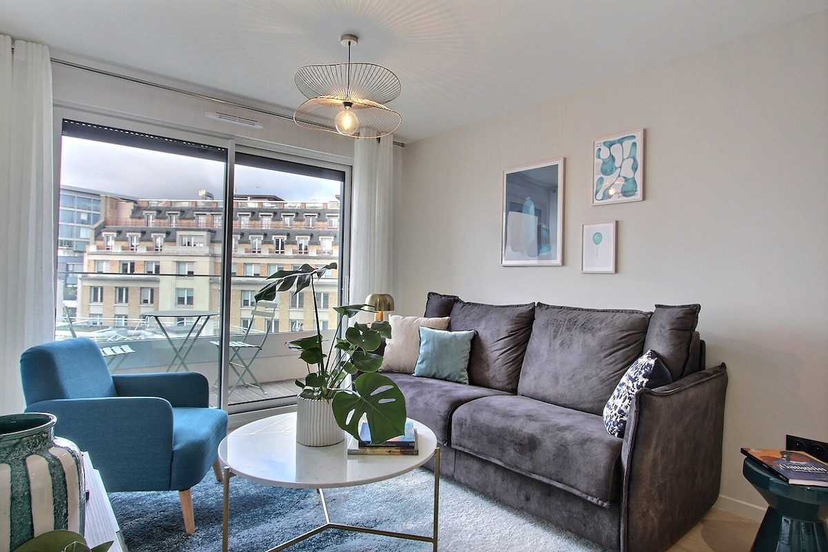 A spacious living area showcases a comfortable gray sofa paired with a blue accent chair. A round coffee table sits in the center, adorned with a small plant. Large glass doors offer access to a balcony, providing natural light and a view of the surrounding architecture.