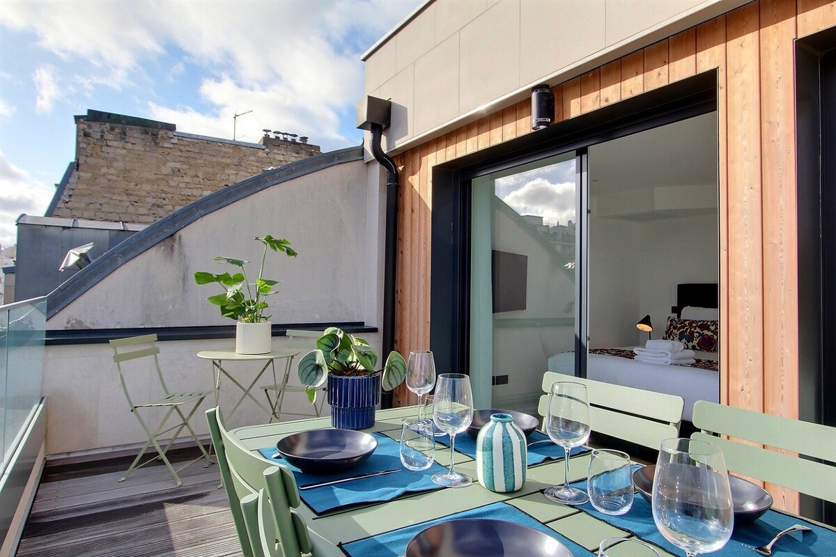 A private terrace is showcased with a set of dining tables and chairs, inviting outdoor meals. A round plant pot sits on the table, while a vase with a striped design adds a colorful accent. The background features a glimpse of the interior through glass doors.