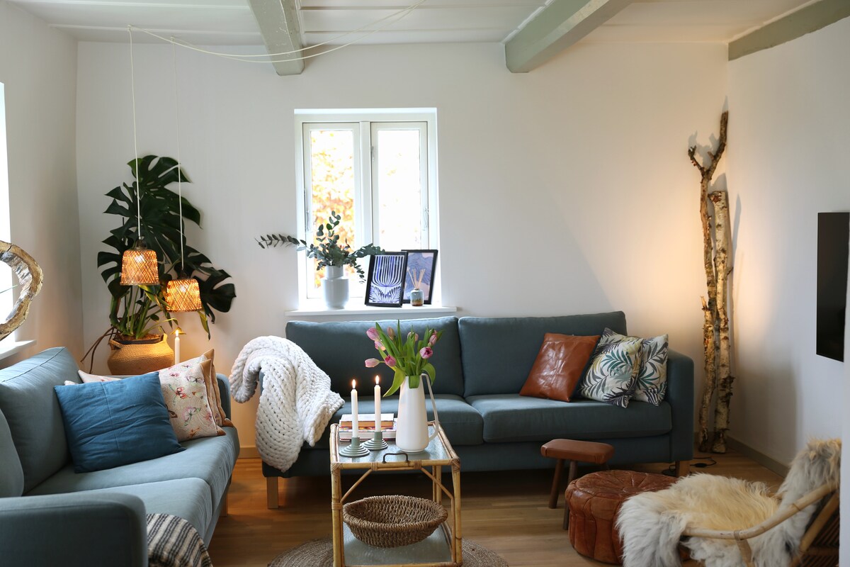 A light-filled living room features two comfortable sofas and a cozy armchair adorned with a soft throw. A coffee table displays fresh flowers, and warm lighting fixtures enhance the inviting atmosphere. Natural elements, including potted plants and decorative wood accents, are present, providing a homely touch.