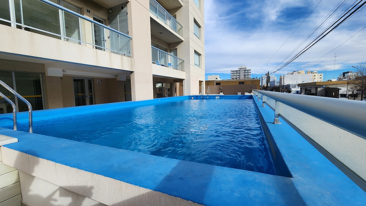 A large outdoor pool is framed by a bright blue perimeter, reflecting the clear sky above. The pool area is surrounded by modern buildings, with lounge chairs visible at the water's edge, providing a serene space for relaxation.