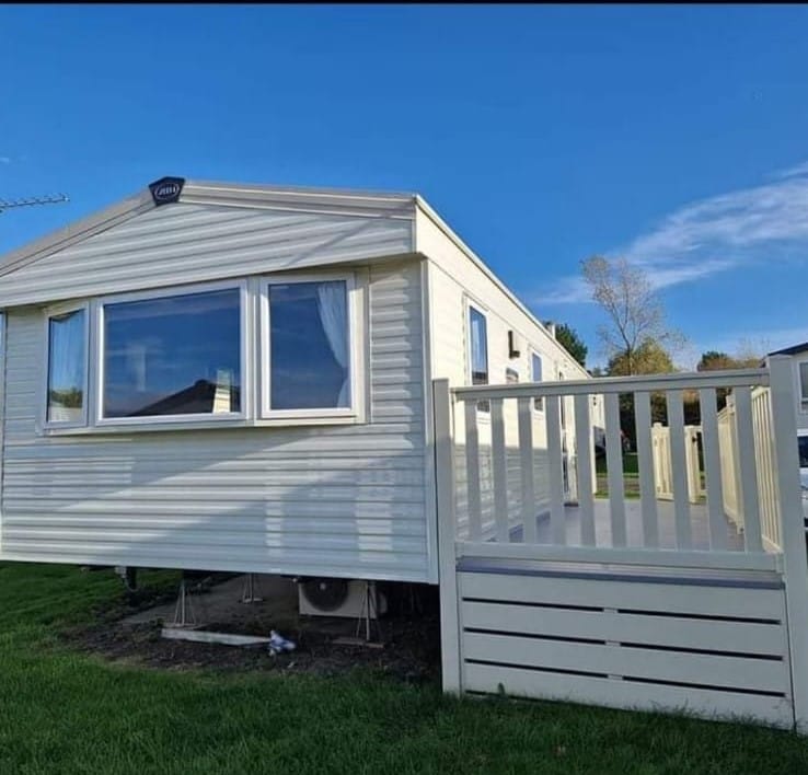 The modern caravan is showcased against a clear blue sky, featuring a spacious deck with a railing. A large window provides an unobstructed view of the interior, and the exterior is painted in soft hues, blending with the surrounding green landscape.
