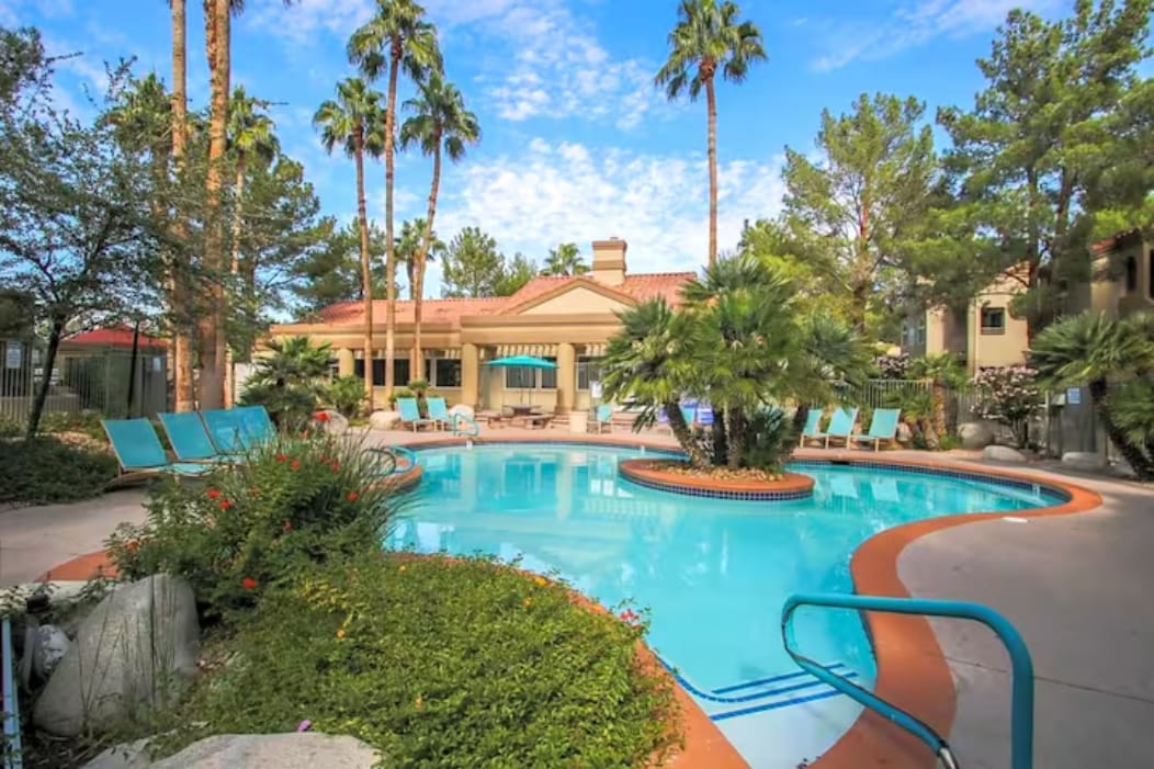A clear blue swimming pool is surrounded by palm trees, vibrant bushes, and lounge chairs. A welcoming building with a green awning is visible in the background. The area is sunlit, showcasing a relaxing outdoor environment.