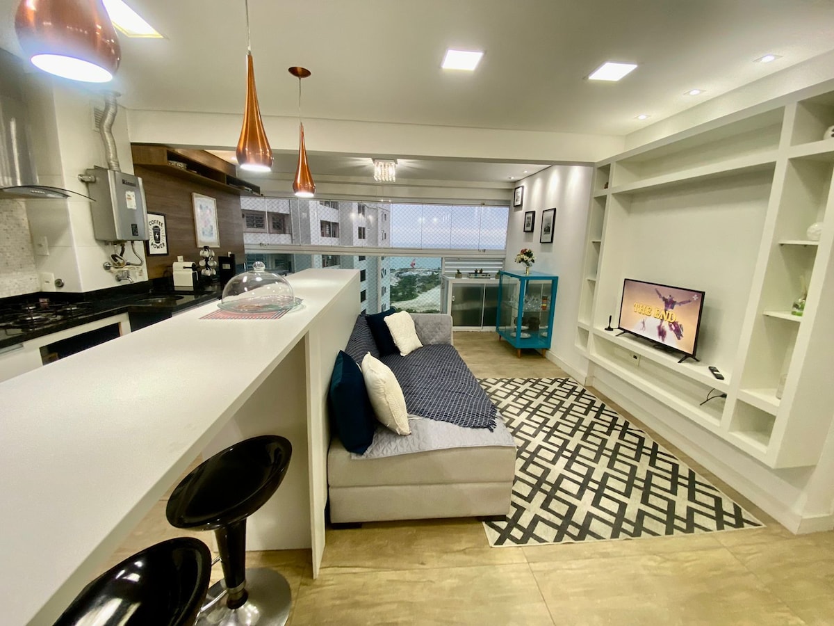A modern living area features a spacious gray sofa adorned with white and blue pillows, positioned in front of a sleek TV unit. Natural light floods the space through large windows, showcasing a charming view. A stylish rug adds depth to the contemporary design.