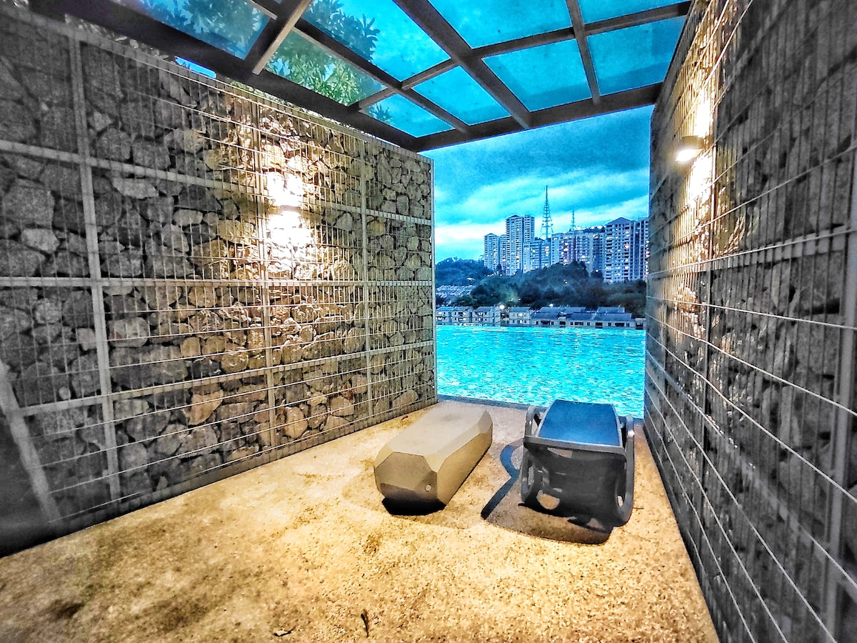 A shaded outdoor area features stone walls and ambient light fixtures. Two lounge chairs are positioned facing a body of water, creating a comfortable space for relaxation while enjoying views of the city skyline beyond.