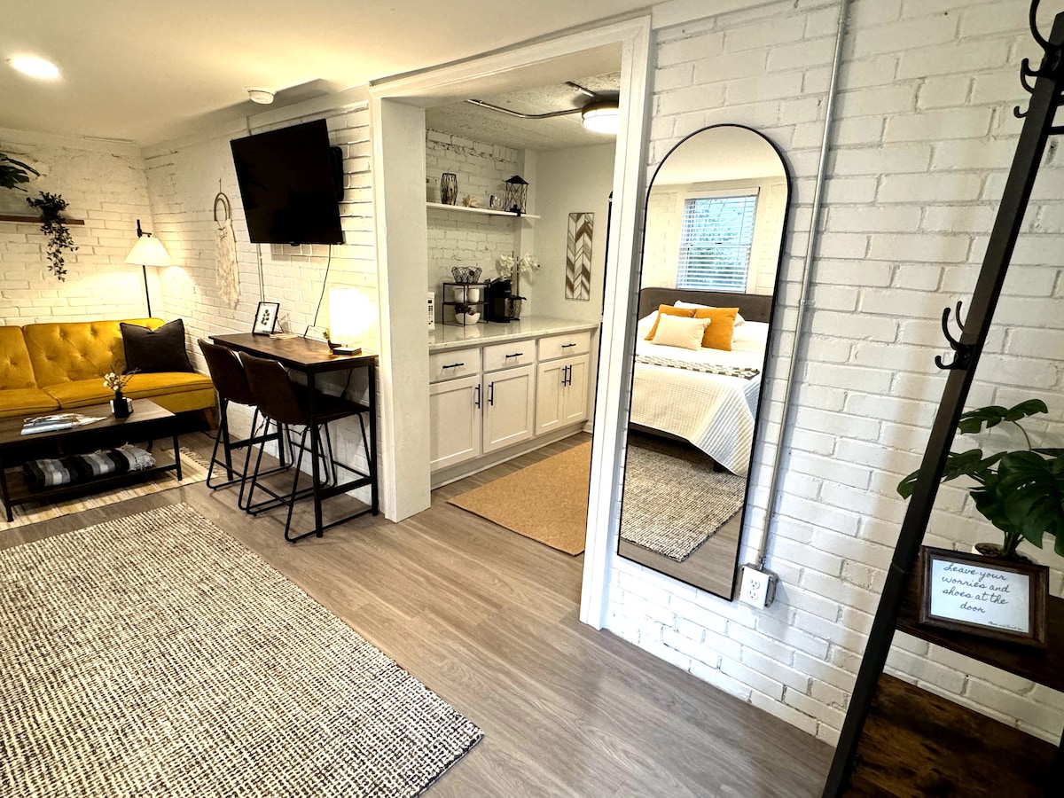 The interior features a neutral color palette with white brick walls. A cozy seating area includes a yellow sofa and a dining space with two stools. A mirror and a mounted TV are visible, with a kitchenette area seen in the background, enhancing the modern design.