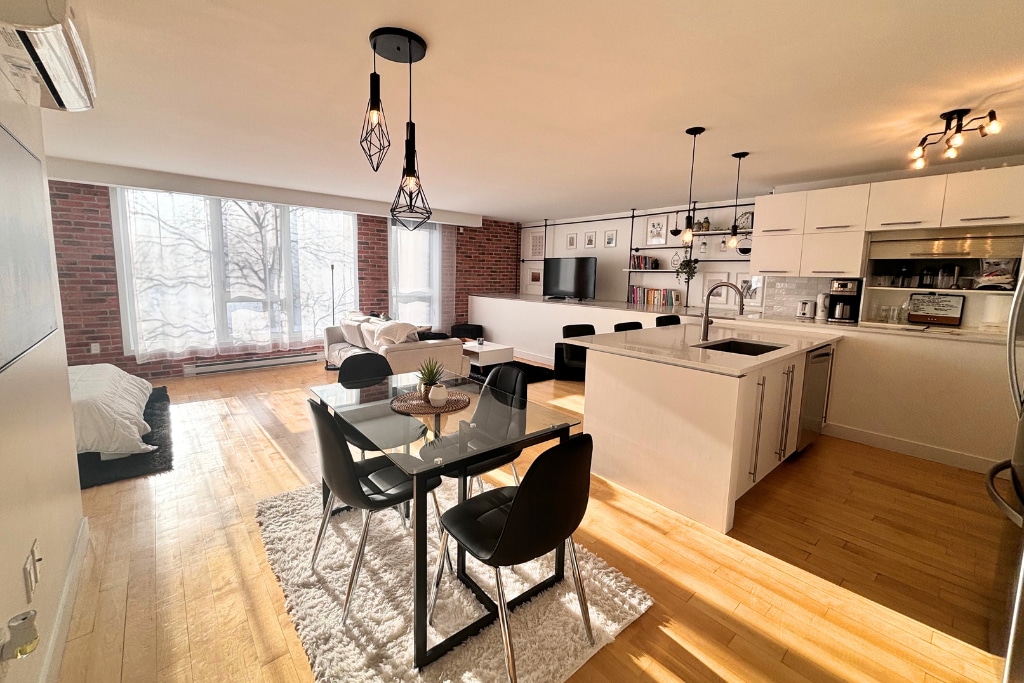 An open-concept living area is visible, featuring hardwood floors and exposed brick walls. A dining table with four chairs sits in the foreground, while a spacious lounge area with floor-to-ceiling windows allows ample natural light. A modern kitchen is equipped with quartz countertops.