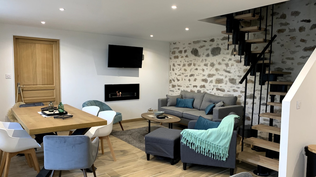 A modern living space is presented, featuring a light wood dining table surrounded by white and blue chairs. A comfortable gray sectional sofa faces a mounted television, with a staircase leading to a loft area. Warm lighting highlights the textured stone wall.