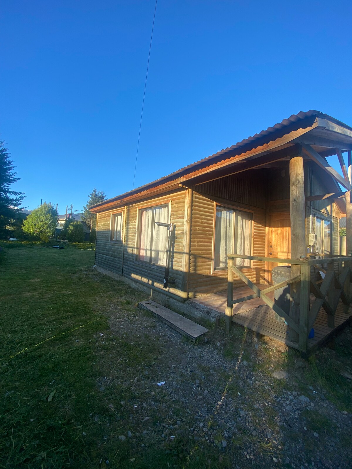 The exterior of a charming wooden cabin is highlighted, featuring a welcoming porch with steps leading to the entrance. Surrounded by greenery, the cabin's natural wood finish blends with the tranquil setting under a clear blue sky.